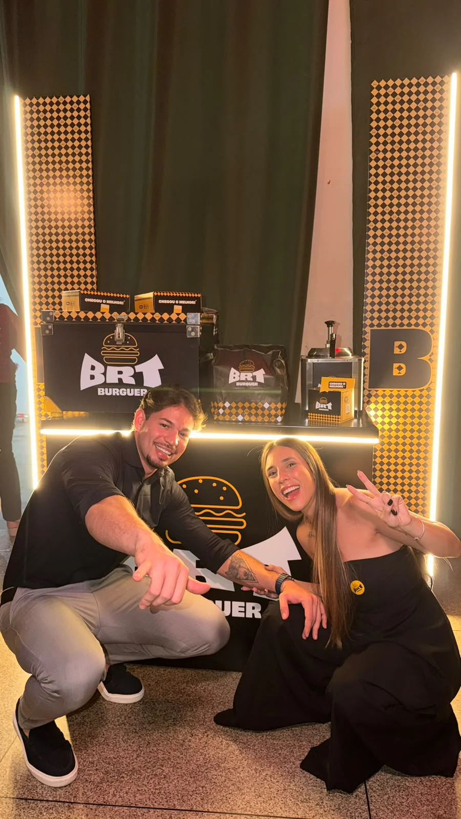 Gabriel e Geovanna, donos do BRT, agachados e sorrindo diante do balcão decorado com o logotipo da marca e o quadriculado amarelo e preto