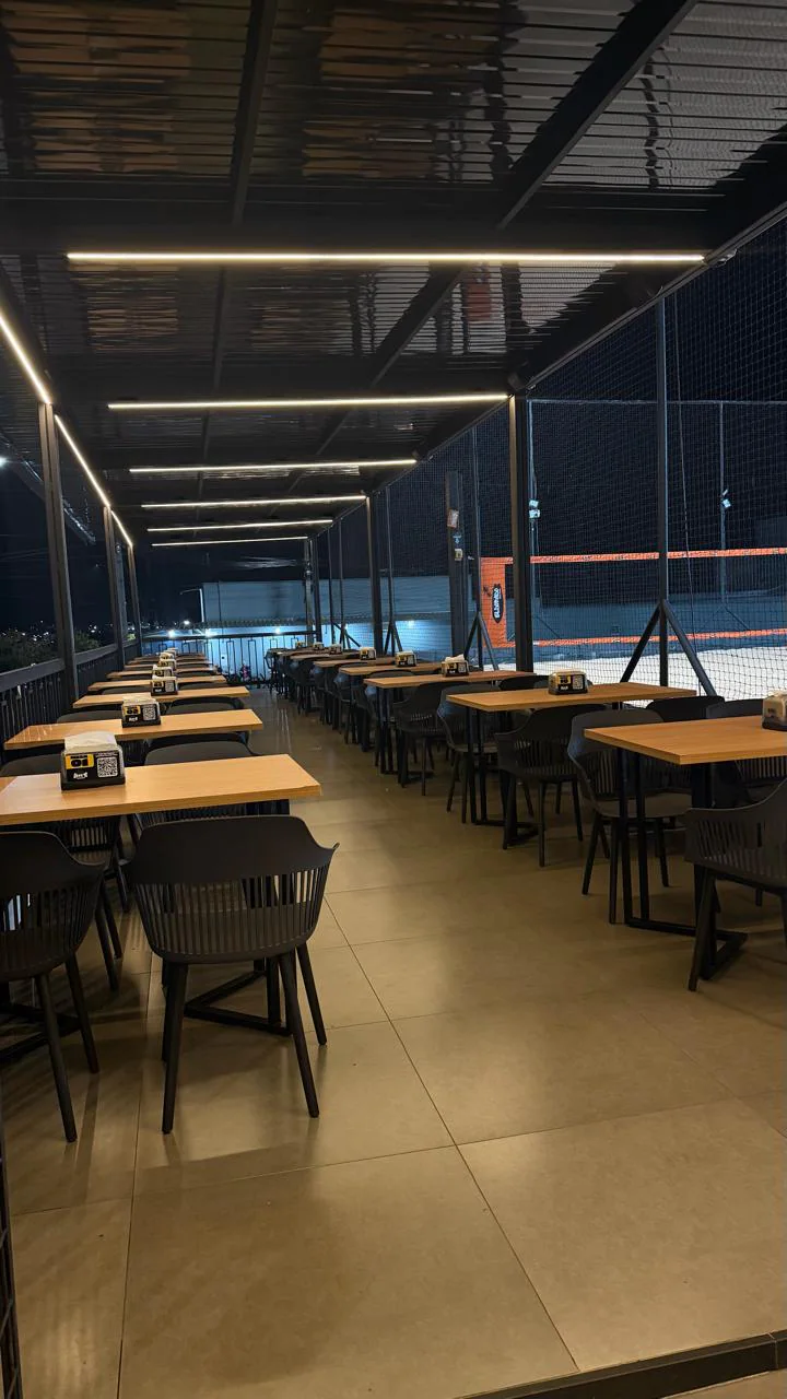 Fileiras de mesas de madeira com cadeiras pretas sob a cobertura iluminada por frisos de luz, ao lado de uma quadra de areia à noite